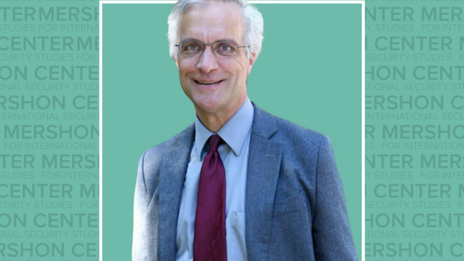 An image of a man with gray hair wearing glasses and a suit, standing before a green background with the Mershon Center logo pattern