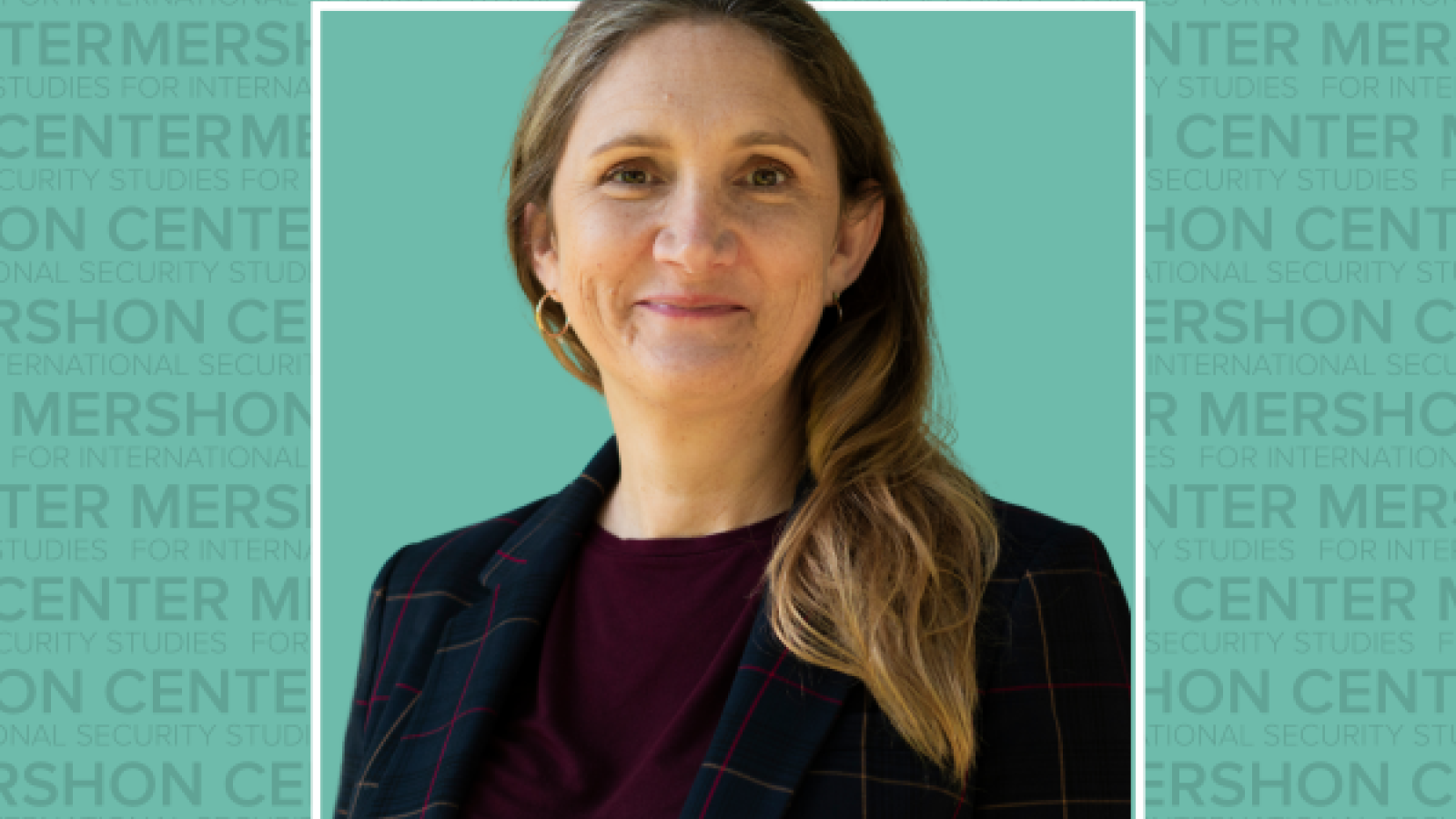 A photo of a woman with shoulder-length hair and a blazer, smiling in front of a turquoise background with the Mershon Center logo.