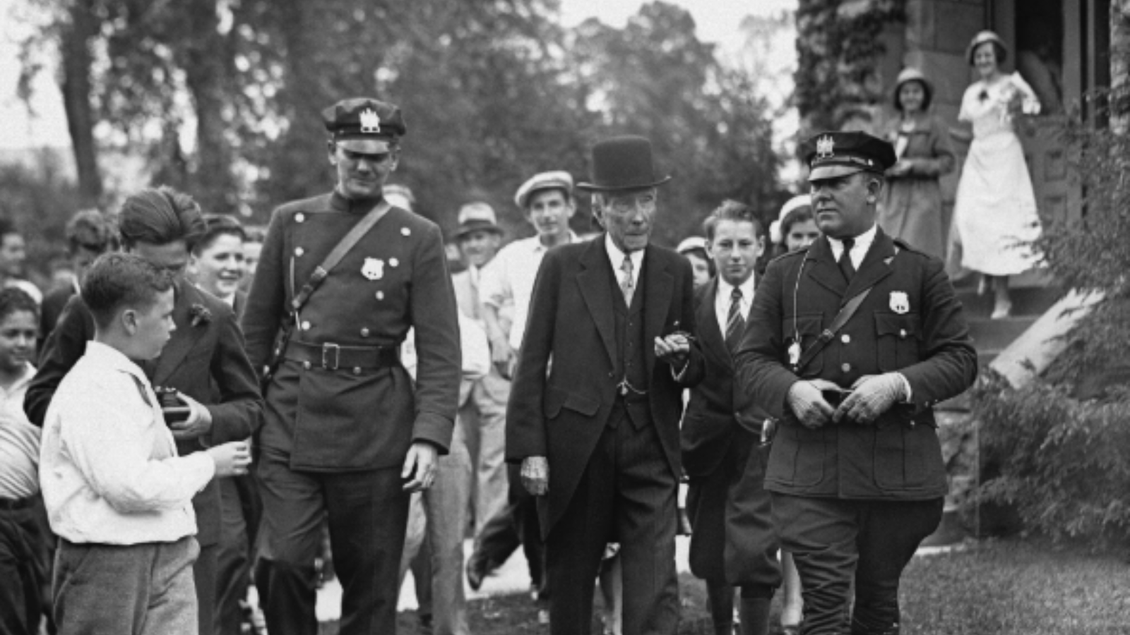 John Rockefeller surrounded by people and flanked by police officers